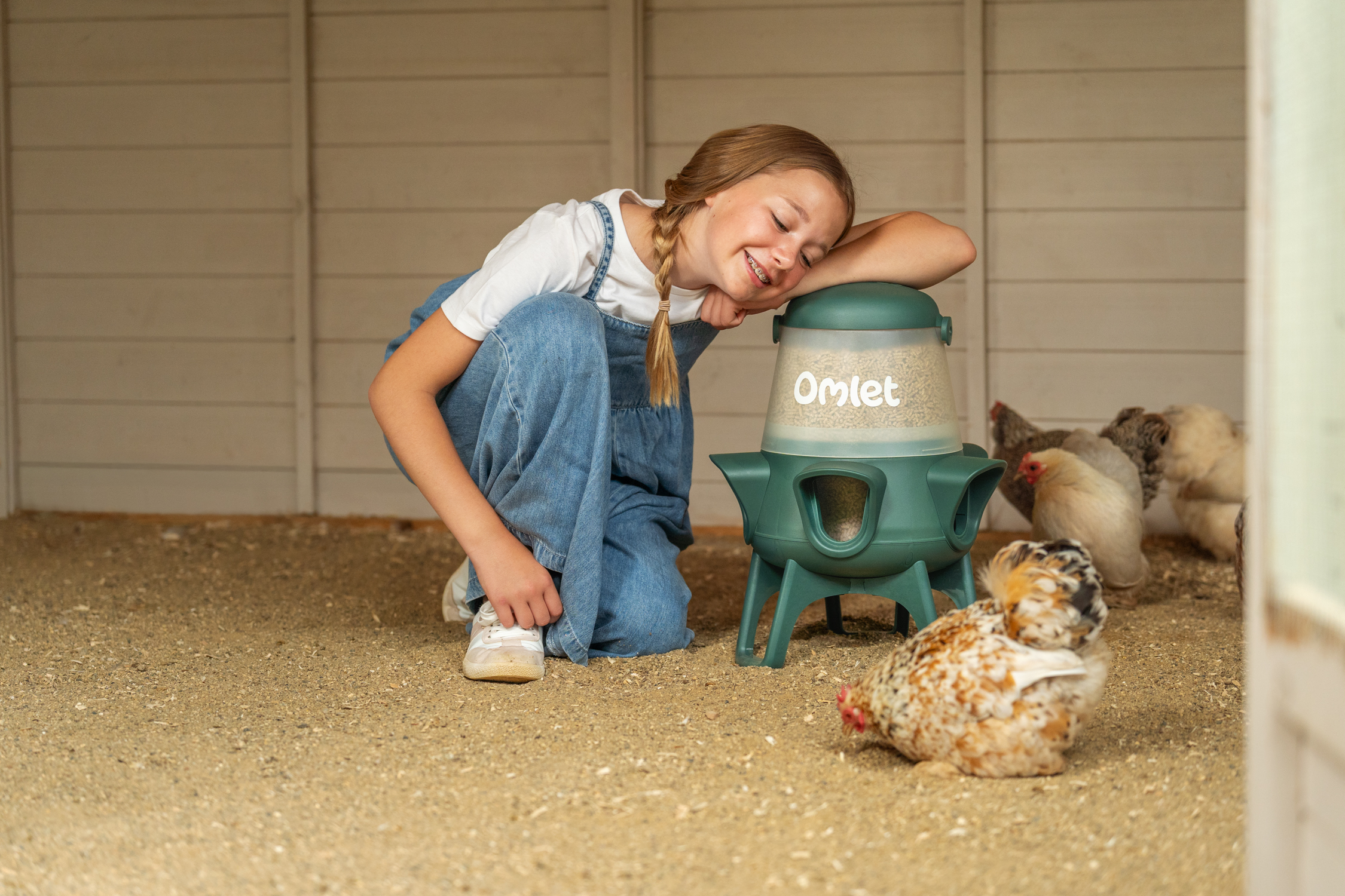 girl leaning on no waste chicken feeder with chickens scratching around a wooden chicken coop